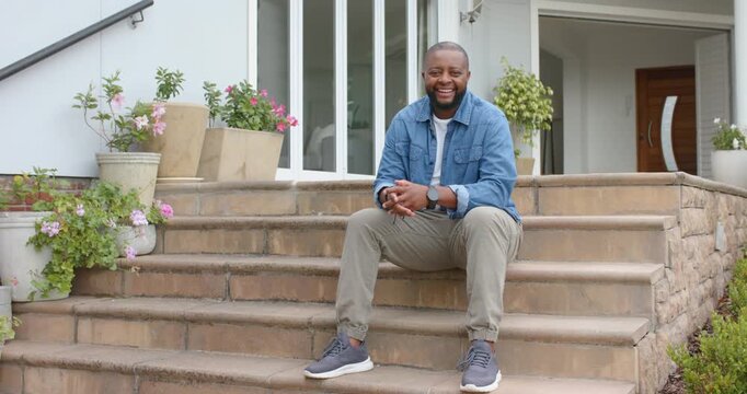 Adult African man wearing mic responding to off-camera prompt, shifting on stone steps and laughing