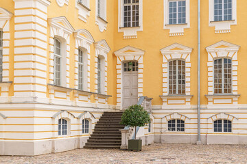 Warm yellow walls frame classic white trim and tall windows. Stone steps lead to a sturdy wooden door, flanked by potted greenery