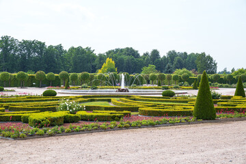 Obraz premium Symmetrical garden with manicured hedges and fountain centerpiece. Greenery frames tranquil water feature under clear sky. Tall trees offer background contrast to geometric designs