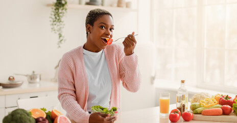 A woman stands in a sunny kitchen holding a bowl of salad and a spoon. She smiles as she takes a bite of the food. Fresh vegetables and a glass of juice are visible on the table. © Prostock-studio