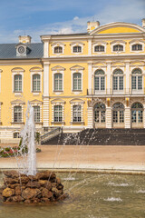 Grand yellow Rundale palace with classical architecture. Fountain jets water in center of stone pond. Symmetrical gardens frame the elegant structure. Sunny sky enhances the scene's calm elegance