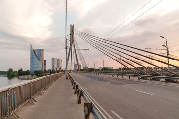 Sunset paints sky soft orange and pink Modern cable-stayed bridge spans calm river waters in Riga. Steel cables stretch from tall central tower Urban skyline looms behind, with glass and steel towers