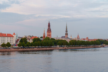 Fototapeta premium Warm sunset bathes historic buildings in Riga Latvia beside calm river Duna. Trees line the waterfront, framing the skyline softly. Red-tiled roofs and spires glow in golden light Panorama