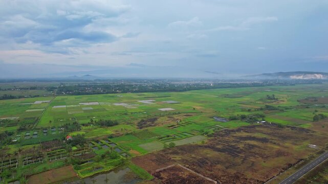 Aerial drone footage of the flat green area in Jalan Lintas Selatan, nearby the coast of Lumajang regency, Java island, Indonesia, with many crops and grass fields, local villages, at cloudy sunset