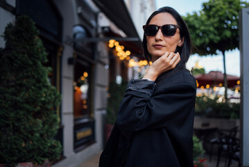 Obraz premium Stylish woman wearing sunglasses poses confidently outdoors, surrounded by greenery and warm ambient lights, showcasing modern fashion and urban lifestyle