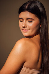 Fototapeta premium Young woman with long dark hair, smiling softly with closed eyes, showcasing natural beauty and serene expression against a warm, neutral background, emphasizing tranquility and self-acceptance