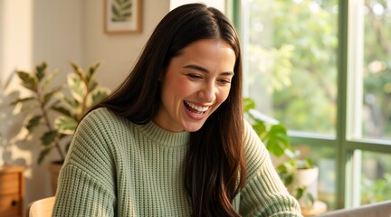Young woman laughing while working on a laptop at home. Happy female student in a green sweater in a bright room with plants. Remote work and lifestyle concept