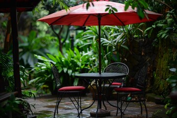 Fototapeta premium Rain falling on a red patio umbrella covering a table and chairs in a lush tropical garden setting