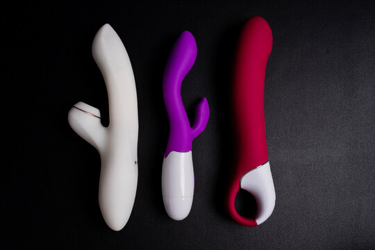 White suction vibrator with clitoral stimulator, purple rabbit vibrator and red phallic vibrator placed on dark textured background under studio lighting.