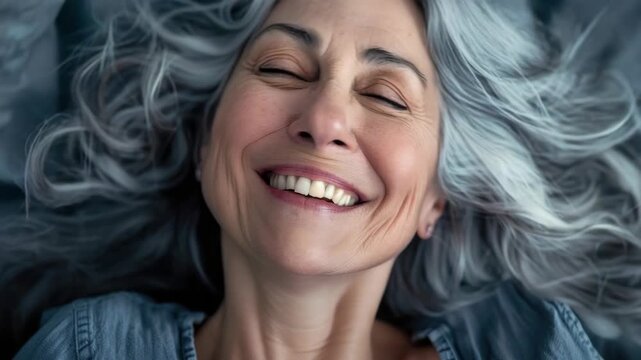 Soft curls frame a joyful, relaxed expression. Eyes gently closed, lips curve in warm, genuine smile. Silver hair flows around a tranquil, contented face. Denim top adds casual, effortless charm