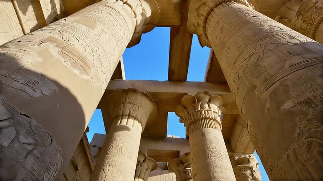 Kom Ombo temple columns with composite capitals and hieroglyphic carvings viewed from below against blue sky in Egypt, upward perspective of ancient sandstone architecture.