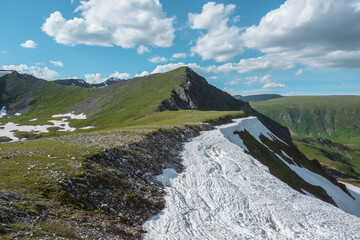 Scenic sunlit view along grassy ridge with white snow cornice to green hill top with rocky sheer crags in bright sun under lush clouds in blue sky. Vivid alpine scenery in high mountains in sunny day. © Daniil