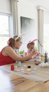 Vertical video: Mother and daughter at table by window, mother teaching egg painting with brushes