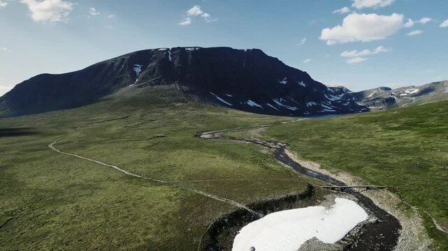 Drone flying up, captures a view of Bl&aring;h&oslash;a mountain in Trollheimen Oppdal Norway. Hikers explore the trail, a bridge crosses the stream, and snow remains on the ground in summer