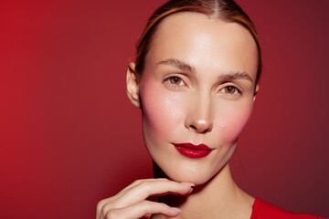Fototapeta premium Close-up portrait of a woman with glossy red lips and pink blush on cheeks against red background. Concept of beauty, makeup, skincare, and fashion style.