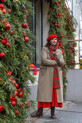 A young beautiful girl in a red beret and beige coat with a bouquet of fir branches walks along the Christmas-decorated streets of the city.