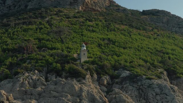Rocky Coastline With Lone Lighthouse Perched On Verdant Mediterranean Hillside, Rugged Rocks In Foreground, Calm Sea, Warm Light, Historic Maritime Beacon Standing