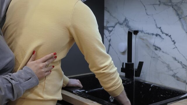 Hands guiding partner cleaning up at kitchen sink, yellow sweater and gray jacket, red nails resting on back, modern marble backsplash and black faucet, turning tap and flowing water, tender support
