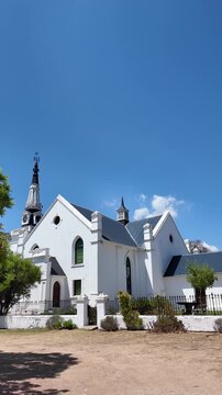 Hopefield South Africa. 10.02.2026. A panoramic view of a classic Dutch Reform church building with a tall spire, surrounded by green trees and a dusty ground on a sunny day in Hopefield South Africa.