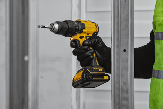 Builder in protective gloves and high visibility vest using cordless drill to install metal stud frame indoor. Concept of renovation, drywall installation, interior construction and professional work.