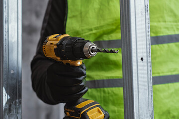 Builder in protective gloves and high visibility vest using cordless drill to install metal stud...