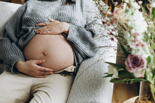 Stylish pregnant woman hugging baby bump close up while sitting on sofa near bouquet. Happy Mothers day! Healthy pregnancy and fertility concept.