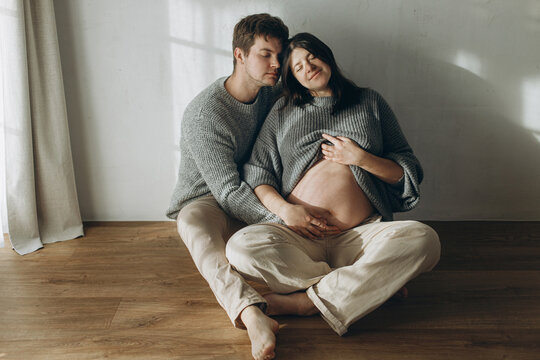 Stylish pregnant couple hugging baby bump and sitting on floor. Fertility, healthy pregnancy and expecting new life concept. Happy Family in cozy sweaters and jeans holding pregnant belly