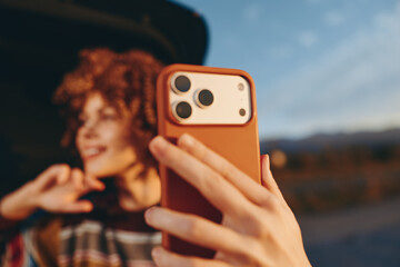 A woman with curly hair wearing a rainbow sweater smiles while taking a selfie with a smartphone outdoors during golden hour. The blurred background shows a natural landscape.