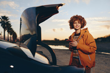 Young woman smiling outdoors near car trunk at sunset holding smartphone wearing rainbow sweater...