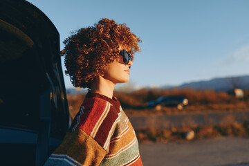 Woman with curly hair wearing sunglasses and rainbow sweater stands outdoors near car trunk,...
