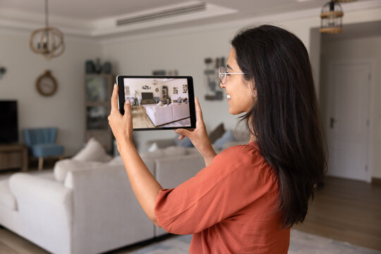 Side view of smiling designer or realtor woman using digital tablet to take photos or videos of modern furnished living room interior, capturing details for real estate, design and renovation purposes