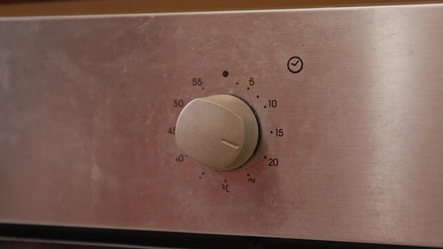 Close up of hand in red sleeve turning a timer knob on stainless steel appliance panel with minute markings and clock icon, adjusting cooking time with shallow depth of field and reflections on metal