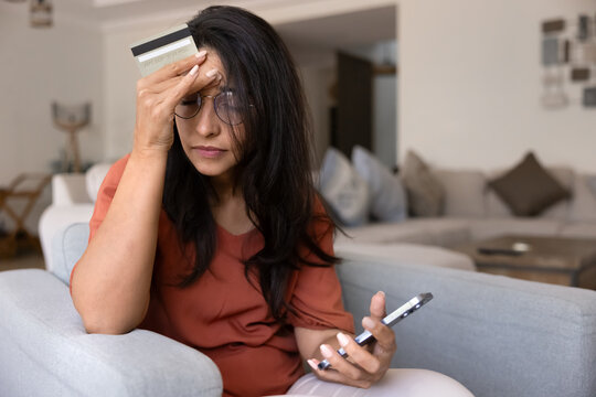Young stressed woman sitting at home with credit card and smartphone, looks concerned or anxious due to online payment failure, financial issues, fraud, digital banking error, unsuccessful transaction