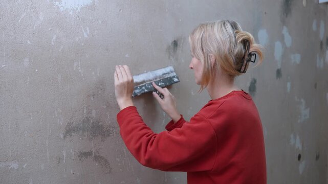Young blonde woman applying plaster with a putty knife, leveling the concrete surface during an independent home renovation project to prepare the wall for painting and decoration