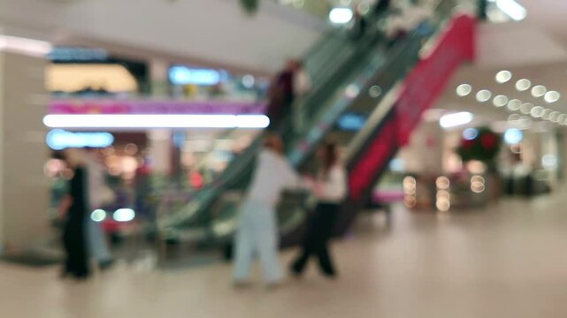 Blurred bokeh background footage of a modern shopping mall interior, with anonymous visitors walking through common areas and moving on an automated escalator connecting different floors.