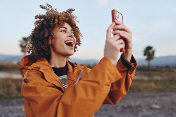 A happy woman with curly hair wearing an orange jacket smiles broadly while holding a smartphone outdoors during sunset. The woman enjoys capturing a moment in nature.