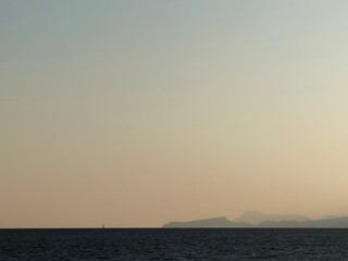Fototapeta premium Minimalist landscape composition with a narrow strip of sea at the bottom edge. Sailboat silhouette on horizon line. Mountains. A vast expanse of soft gradient sky during sunset. Calm and serene.