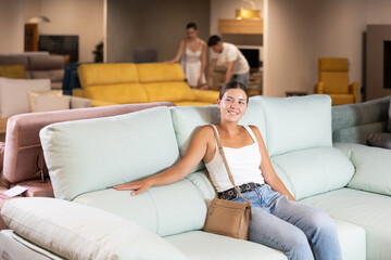 Obraz premium Pleased young girl choosing convenient sofa in large furniture shop