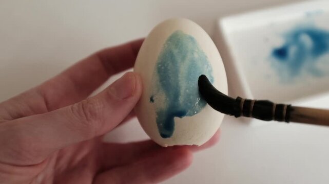 Close-up of the process of dyeing large goose eggs with blue dye, showing hands coloring Easter eggs for spring holiday decorations and traditional DIY family crafts.