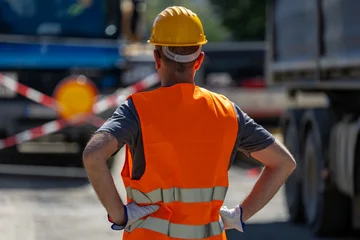 Construction worker stands with his back to the camera wearing a hard hat and safety vest at a road work site in daylight hours © zphoto83