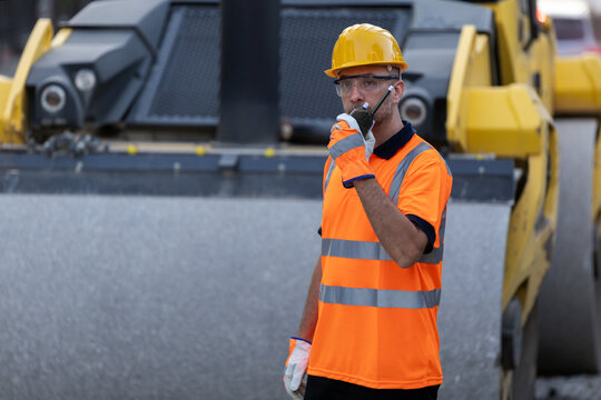 Construction worker in safety gear talks on radio near road construction site with heavy machinery during the day
