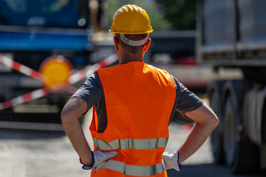 Construction worker stands with his back to the camera wearing a hard hat and safety vest at a road work site in daylight hours