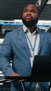 Vertical video Server farm programmer at workplace desk using laptop to optimize electronics. African american man in data center using notebook to monitor system performance, identifying bottlenecks