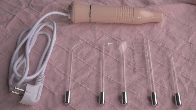 Skincare device with glass attachments and power cord on soft pink fabric background for beauty treatment. New electric Darsonval. Skincare woman concept
