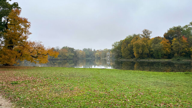 Scenic wide-angle view of Apollo Temple (Monopteros) across a misty lake in Nymphenburg Palace gardens, Munich, during autumn.