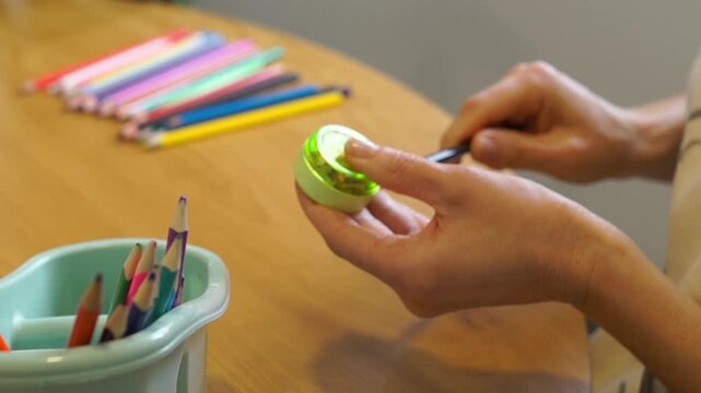 A woman takes different colored pencils from a container and sharpens them one by one using a green sharpener on a wooden workspace.