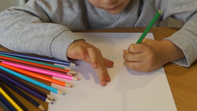 A young boy writes with a green pencil using his left hand, looks at the broken tip, and prepares to continue his work.