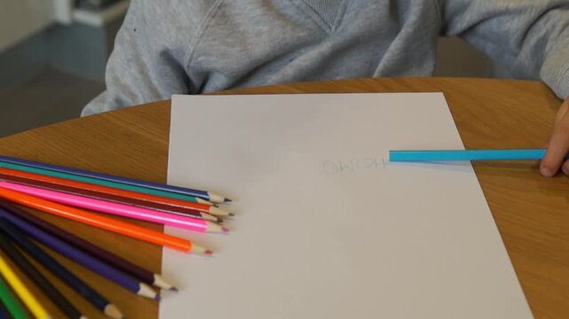 A left-handed child focuses on writing the word "HOME" in blue colored pencil, with a variety of crayons scattered on the desk.