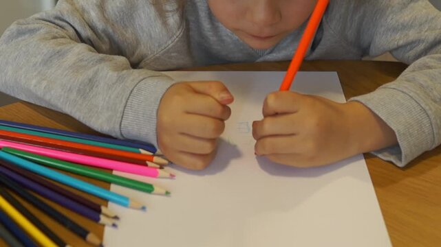 Close-up of a left-handed boy carefully writing on a white sheet of paper using an orange colored pencil, surrounded by school supplies.