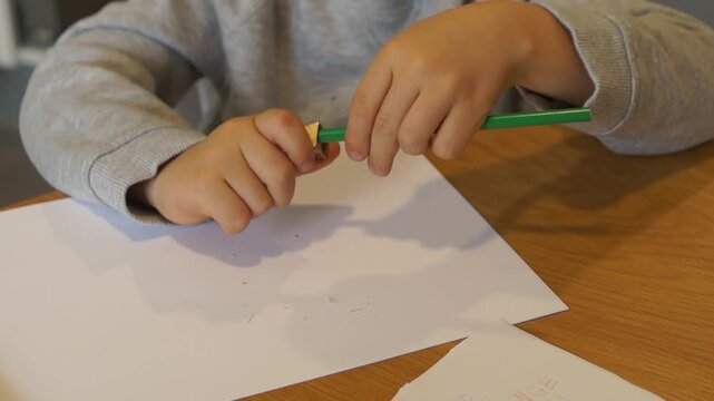 A close-up of a young left-handed boy carefully using a metal sharpener to sharpen a green colored pencil over a white sheet of paper.